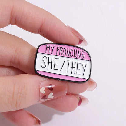 Pins pronoms She/They tenu entre deux doigts